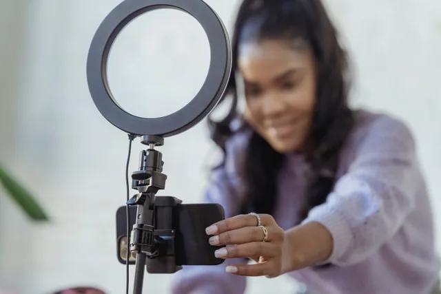 Cheerful young African American female blogger in stylish sweater smiling while setting up camera of smartphone attached to tripod with ring light before recording vlog. Real Estate Social Media: A Realtor's Process Guide