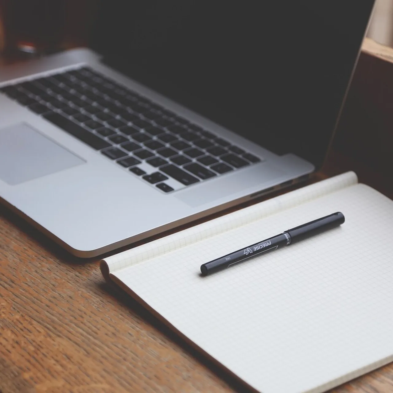 Open laptop beside a blank grid notebook and black pen on a wooden desk.